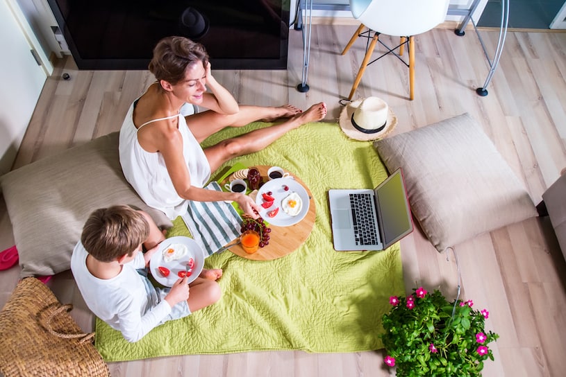 uma mulher e uma criança sentados sobre um tapete e comendo várias frutas enquanto assistem alguma coisa no computador, a ambientação tem uma cadeira, algumas almofadas, um chapéu de palha, um arranjo floral e uma bolsa de sisal