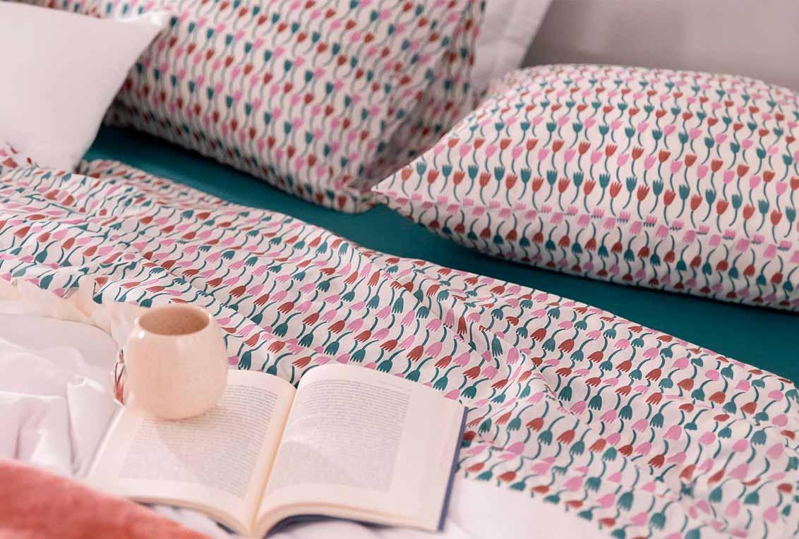Conjunto de fronha e lençol estampados na cama posta. No centro da cama estão um livro e uma xícara redonda.