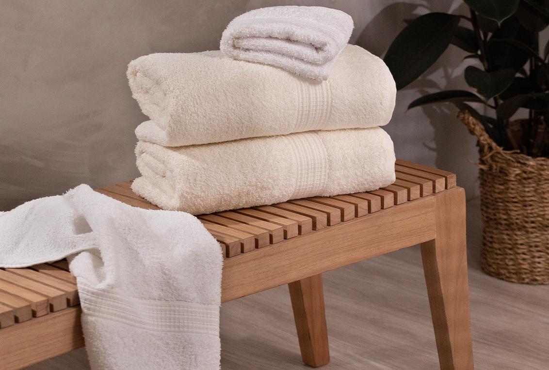 Na imagem está centralizada toalhas de cores diferentes sobre um banco de madeira. Três toalhas estão dobradas, enquanto uma está aberta e posta sobre a superfície do banco.